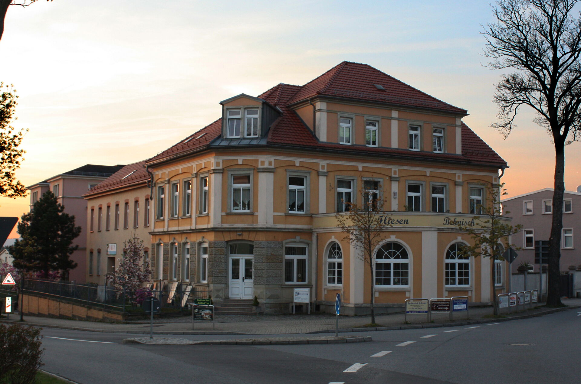 Das gelbe Eckgebäude des Fliesenfachbetriebs Behnisch GmbH in Pulsnitz bei Abenddämmerung. Straße und Bäume im Vordergrund.