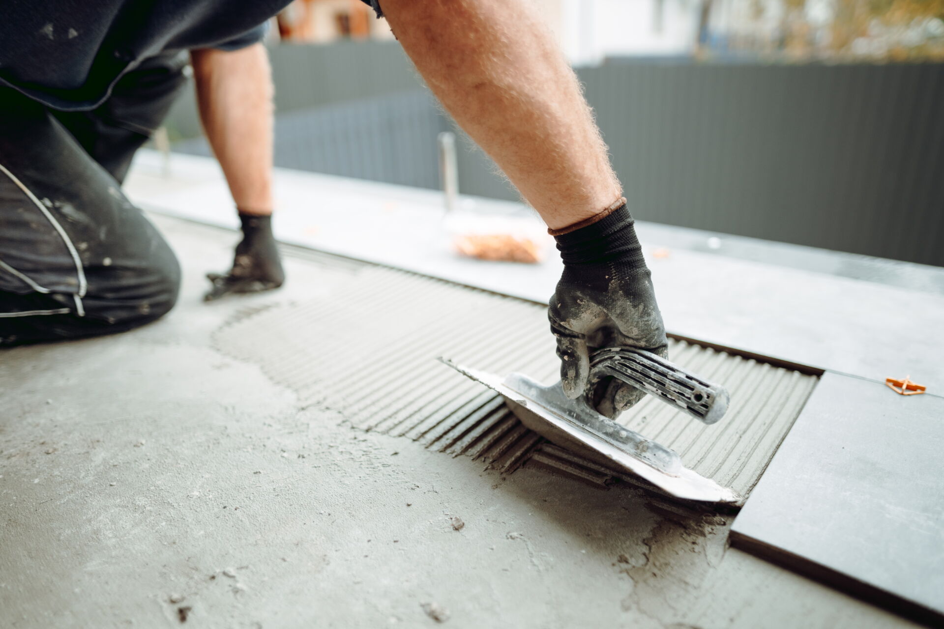Ein Fliesenleger mit Handschuhen verlegt sorgfältig große, graue Fliesen mit einer Kelle. Arbeit des Fliesenfachbetriebs Behnisch GmbH in Pulsnitz.