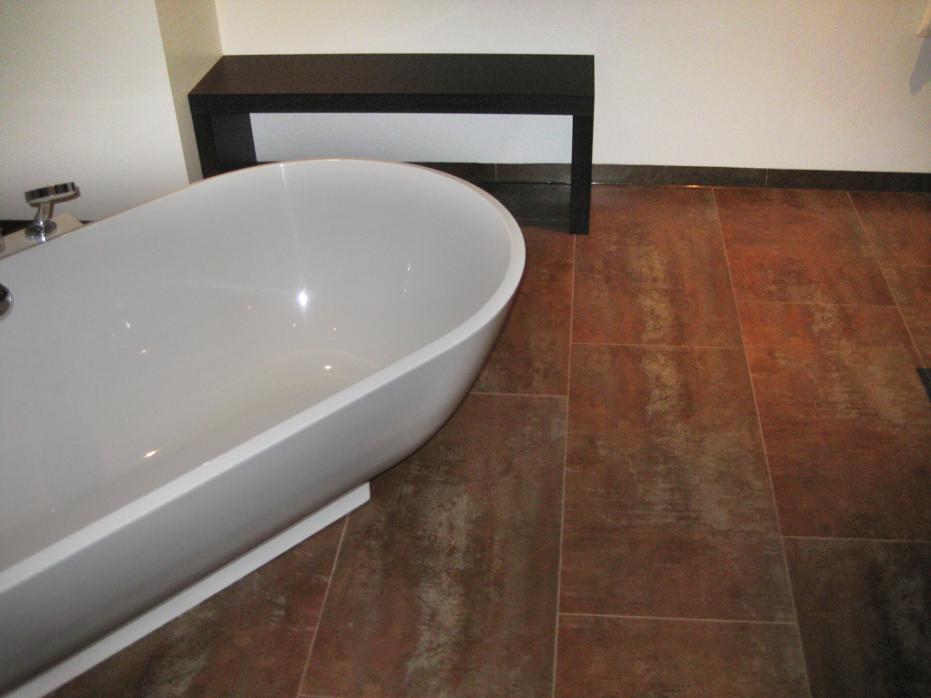 Ein Badezimmer mit einem modern gefliesten Boden in warmen Brauntönen, gestaltet vom Fliesenfachbetrieb Behnisch GmbH in Pulsnitz. Eine weiße Badewanne steht links.