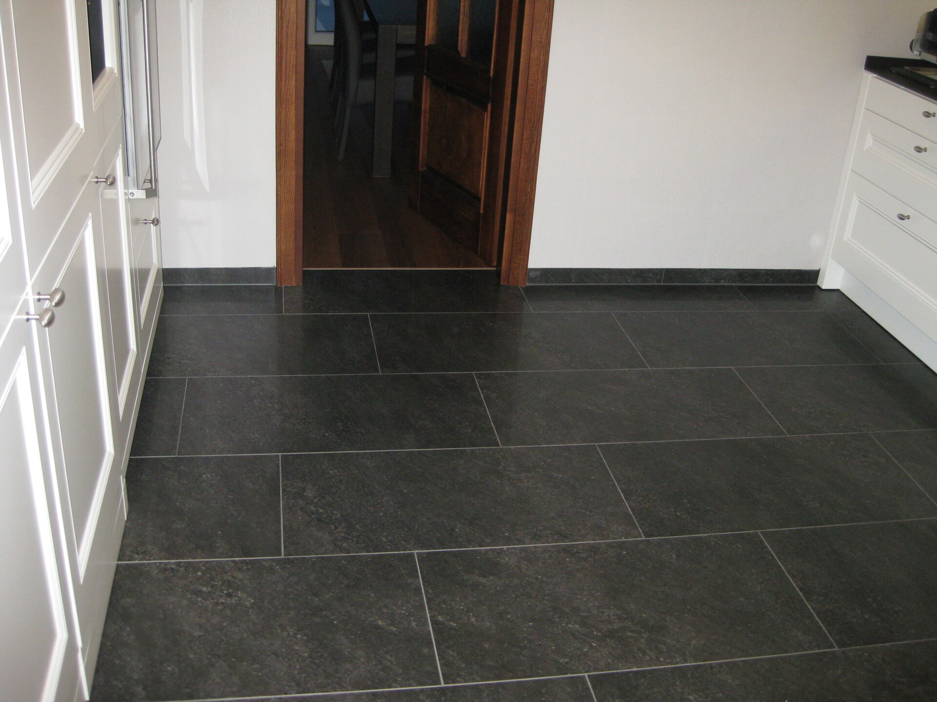 Ein modern gefliester Küchenboden mit großen, anthrazitfarbenen Fliesen, verlegt vom Fliesenfachbetrieb Behnisch GmbH in Pulsnitz. Weiße Küchenmöbel sind sichtbar.