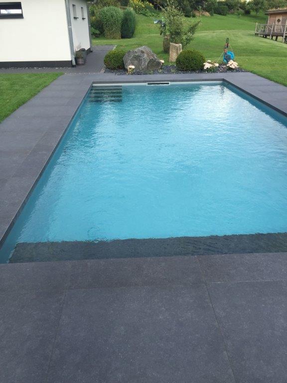 Ein modernes Schwimmbad mit anthrazitfarbenen Fliesen umrandet, eingebettet in einen grünen Garten. Gestaltung vom Fliesenfachbetrieb Behnisch GmbH.